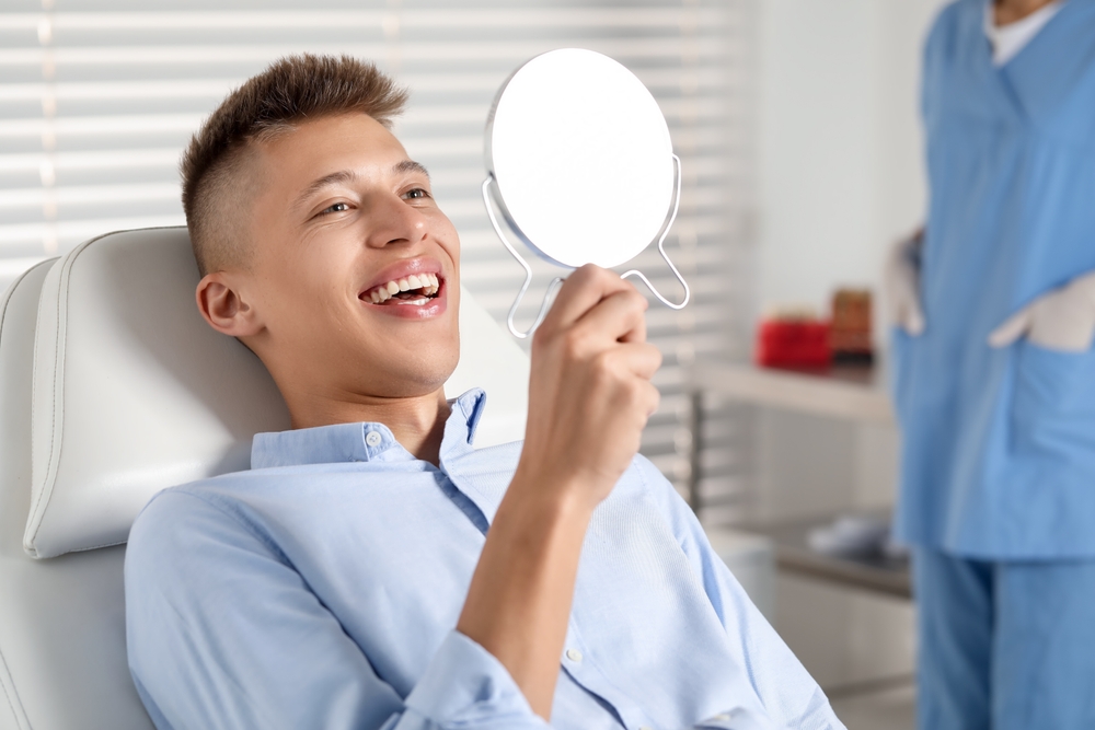 Male patient holding mirror and smiling after successful dental crown restoration – Dental Crowns Highland Park