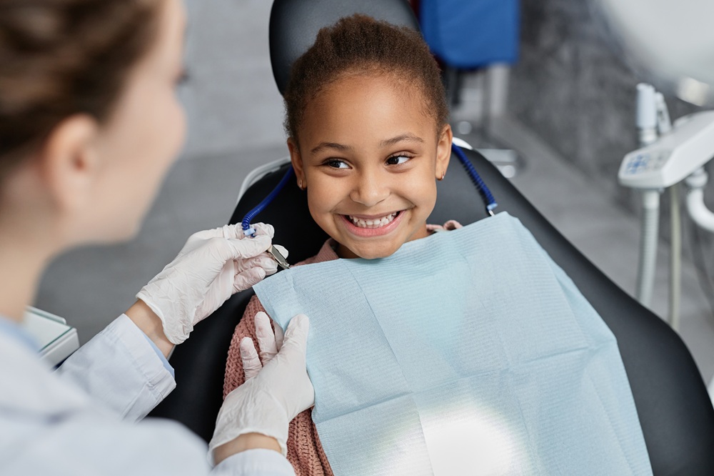 Happy Child at the Dentist - Dentist St Paul MN A young girl with a big smile sits in a dental chair, wearing a protective bib, as a dentist in gloves prepares to examine her teeth. - Dentist St Paul MN